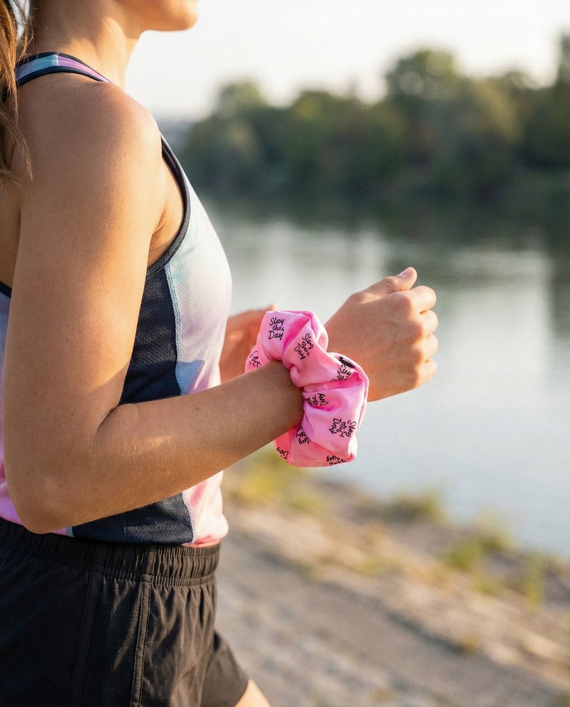 Frau beim Laufen am Flussufer im Morgenlicht, das Slay-the-day-Scrunchie geschlossen als Armband am Handgelenk