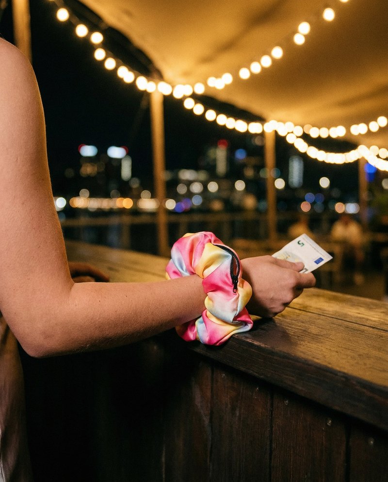 Frauenarm auf einem Bartresen bei Nacht, das Gradient-Scrunchie als Armband am Handgelenk, ein Geldschein in der Hand, Lichterketten im Hintergrund